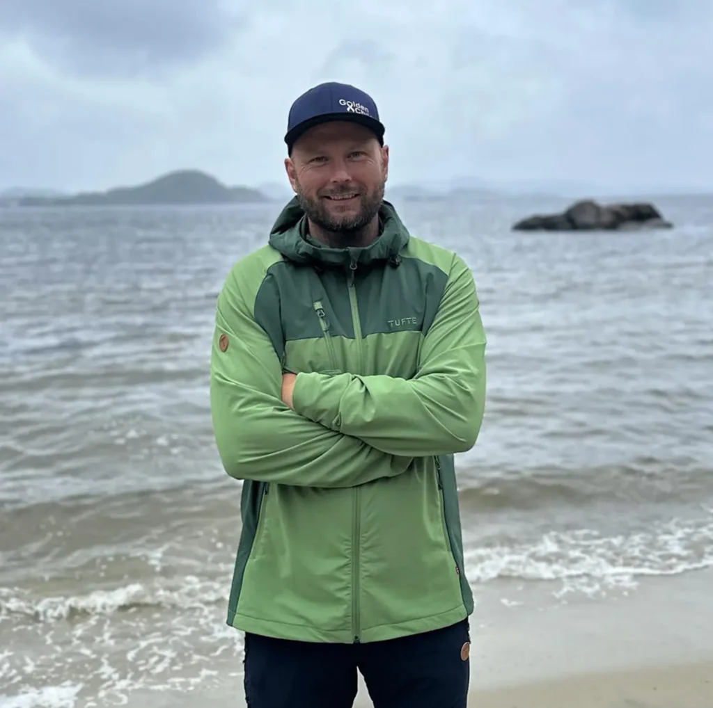 En person iført en grønn jakke og blå caps står med armene i kors på en strand, havet og øyene danner et rolig bakteppe, som om han poserer for et kontakt Tufte Management-reklamebilde.